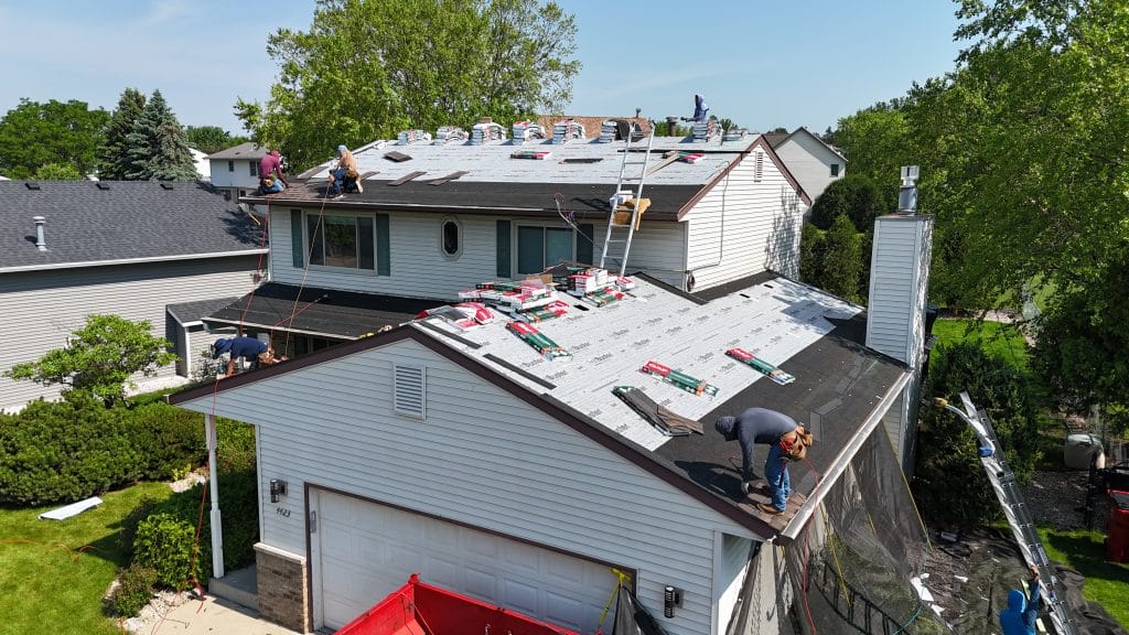 A team of roofers installs new shingles on the roof of a two-story suburban house on a sunny day. Roofing materials are stacked on the roof, and ladders and safety equipment are visible as part of their expert roof repair work.