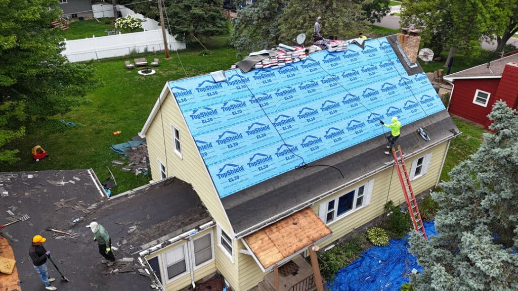 Aerial view of workers from a roofing company installing a new roof on a yellow house. The roof is covered with blue synthetic underlayment, materials are stacked near the chimney, and construction supplies surround the house.
