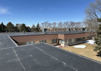 Flat rooftop view of a one-story commercial building with brown brick walls, surrounded by leafless trees and patches of snow on the ground under a clear blue sky.