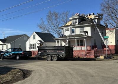 Workers are replacing the roof of a two-story white house on a sunny day. Materials and equipment are on the roof, a ladder leans against the house, and a black truck and trailer are parked nearby.