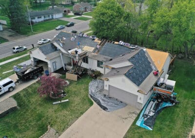 Aerial view of workers replacing the roof on a suburban house, with construction materials and equipment scattered around the property and some parts of the roof stripped and covered with black underlayment.