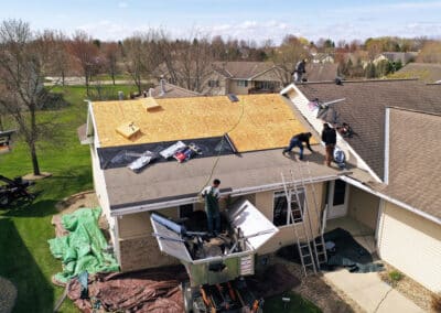 Aerial view of several workers replacing the roof of a house, with some on the roof installing new plywood and others handling materials from a lift below. Tarps cover the ground around the house.