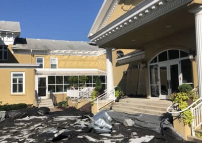 A large yellow building with white columns and arched windows has roofing material and debris scattered on a black tarp covering the ground in front, indicating ongoing roof construction under a clear blue sky.