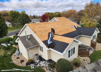 Aerial view of workers installing new plywood sheets on the roof of a beige house during a roofing project; roofing materials and tools are scattered on the lawn below. Trees with autumn foliage are in the background.