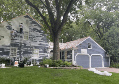 Two people stand on ladders painting the exterior of a blue and white two-story house surrounded by trees. Painting supplies and drop cloths are scattered on the driveway and grass.