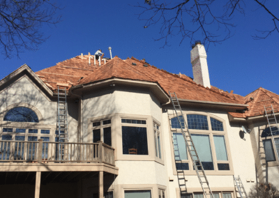 A large house with ladders leaning against it while workers install new wooden shingles on the roof under a clear blue sky. Bare tree branches frame the scene.
