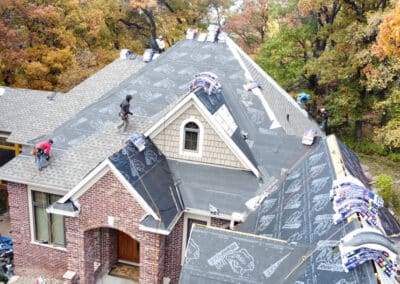 Aerial view of workers installing roofing materials on a large house surrounded by autumn trees. Roofing paper and bundles of shingles are spread across the roof as workers prepare for installation.