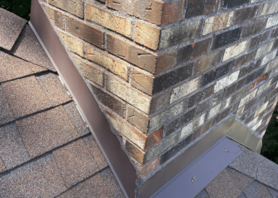 A close-up view of a brick chimney on a shingled roof, showing neatly installed brown metal flashing around the base to prevent water leaks.