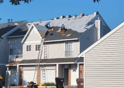 Two workers are repairing the roof of a two-story suburban house using ladders and safety harnesses. Roofing materials are stacked along the roof, and trash bins are placed in the driveway below. It is a sunny day.