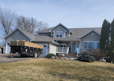 A gray two-story suburban house with a front porch and attached garage, a black trailer parked in the driveway, and a lawn with shrubs and trees in early spring.