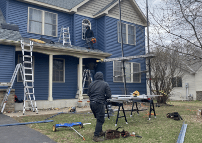 Two workers install or repair siding on a blue house using ladders and scaffolding. Tools and materials are scattered on the lawn, and the sky is cloudy, suggesting an overcast day.