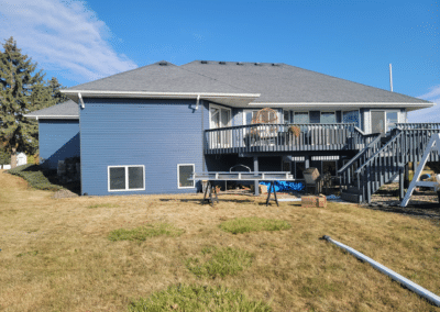 A blue house with a gray roof and raised deck, outdoor furniture, and stairs leading to the lawn. Construction materials and tools are on the grass under clear blue sky.