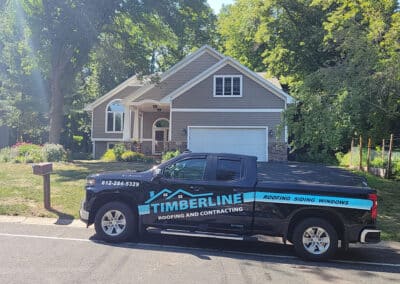 A black Timberline Roofing and Contracting pickup truck is parked on the street in front of a two-story beige house with white trim and a well-kept lawn. Large green trees surround the house and street.