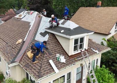 A group of workers in safety gear are installing new shingles and underlayment on the roof of a house, using ladders and various tools. Some areas of the roof are partially stripped, while others are being newly covered.