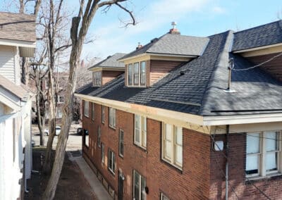 A brick apartment building with dark shingle roofing, dormer windows, and leafless trees along a narrow alley, seen under a clear blue sky.