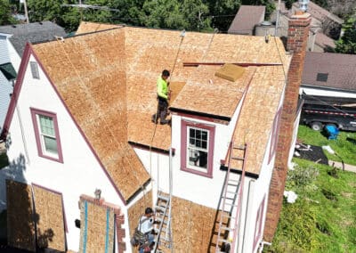 Two workers are installing plywood sheathing on the roof of a house. Ladders lean against the house, and building materials are scattered nearby. The house is surrounded by greenery and neighboring homes.
