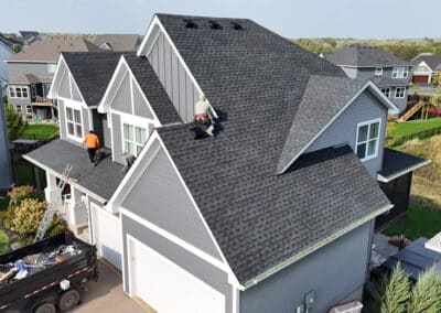 Two workers are installing shingles on the roof of a gray two-story house in a suburban neighborhood. Nearby, a truck with tools and materials is parked in the driveway. Other houses and greenery surround the area.