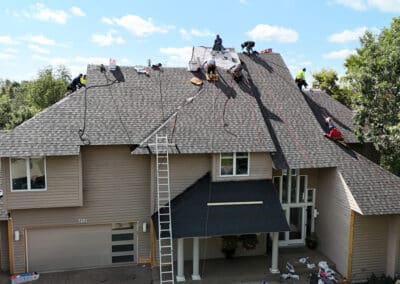 Several workers are installing new shingles on the roof of a large two-story house surrounded by trees, with ladders, tools, and roofing materials visible on the ground and roof.