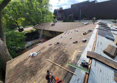 A group of workers repair a large brown shingle roof, with tools, materials, and debris scattered across it. Ladders and a truck are visible below, and trees border the scene on a sunny day.
